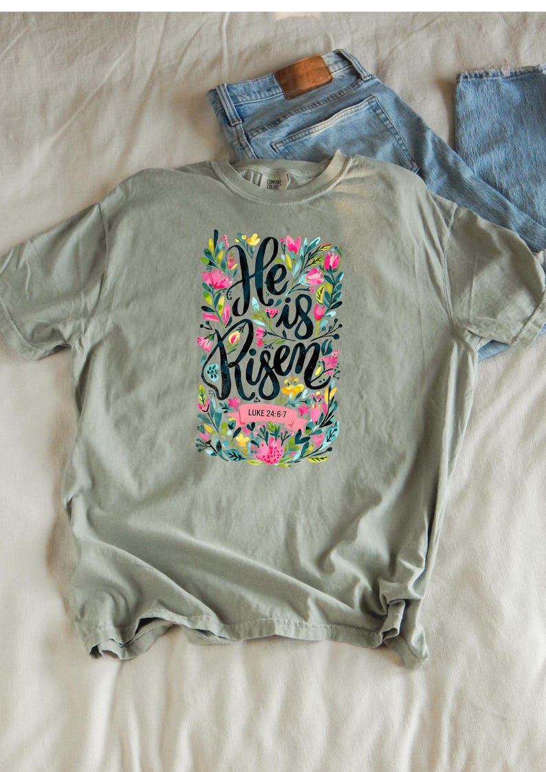 May include: A light green t-shirt with a floral design and the text "He is Risen" with the scripture reference "Luke 24:6-7".