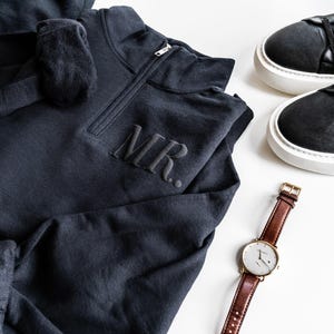 Op de afbeelding: Een donkergrijs sweatshirt met de letters "MR." geborduurd op de borst, gecombineerd met zwarte sneakers en een bruin leren horloge met een goudkleurige kast. Het horloge heeft een witte wijzerplaat.