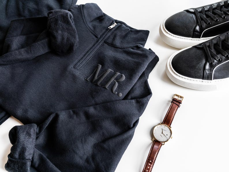 Op de afbeelding: Een donkergrijs sweatshirt met de letters "MR." geborduurd op de borst, gecombineerd met zwarte sneakers en een bruin leren horloge met een goudkleurige kast. Het horloge heeft een witte wijzerplaat.
