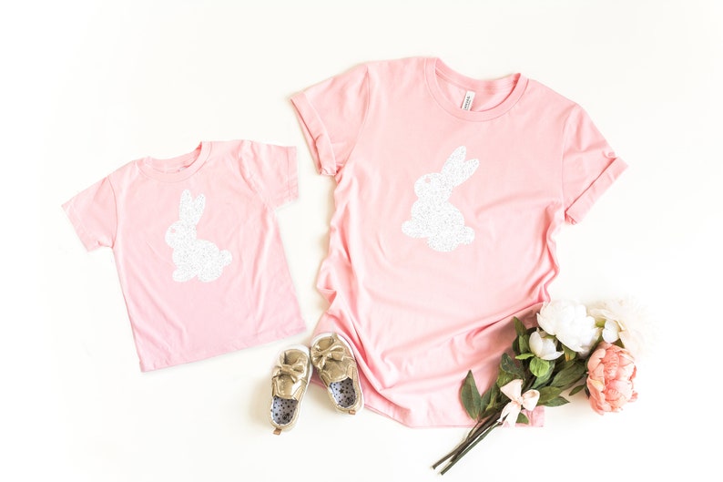 May include: Two pink t-shirts, one adult and one child size, each with a white glitter bunny design. Gold shoes and a bouquet of flowers are also in the image. The shirts are casual wear.