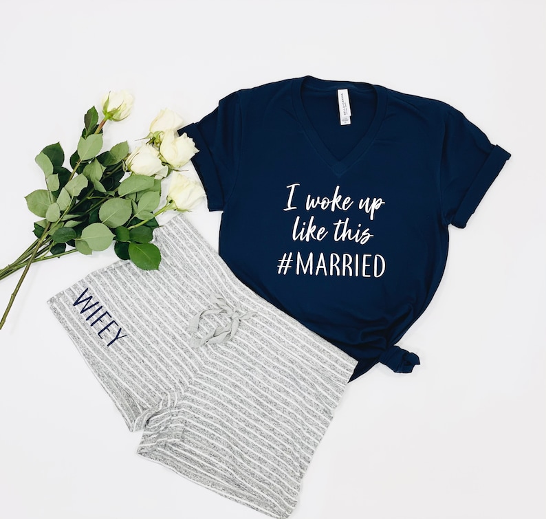 Może przedstawiać: Koszulka w kolorze granatowym z dekoltem w kształcie litery V z napisem "I woke up like this #MARRIED" w kolorze białym. Szorty do piżamy w paski szare i białe z wyszytym napisem "WIFEY" w kolorze niebieskim.