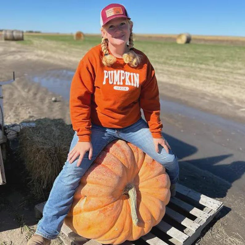 Op de afbeelding: Een oranje sweatshirt met lange mouwen en het woord PUMPKIN in witte blokletters op de borst, gecombineerd met een blauwe spijkerbroek en een bordeauxrode en witte honkbalpet met een rechthoekige patch op het voorpaneel.