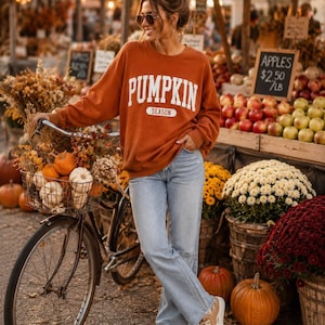 Op de afbeelding: Een roestoranje sweater met ronde hals en de witte tekst PUMPKIN SEASON op de borst, gecombineerd met een lichtblauwe spijkerbroek en beige sneakers. De sweater heeft lange mouwen en een relaxte pasvorm, gedragen in een casual stijl.