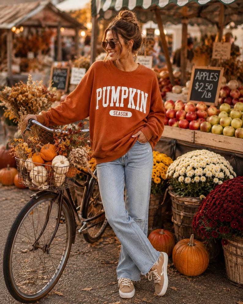 Op de afbeelding: Een roestoranje sweater met ronde hals en de witte tekst PUMPKIN SEASON op de borst, gecombineerd met een lichtblauwe spijkerbroek en beige sneakers. De sweater heeft lange mouwen en een relaxte pasvorm, gedragen in een casual stijl.