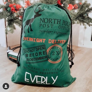 Puede incluir: Una bolsa de regalo navide&ntilde;a verde con cierre de cord&oacute;n negro. La bolsa presenta una silueta de reno y el texto "North Pole Post Office" y "Overnight Delivery". El nombre "Everly" est&aacute; impreso en blanco.