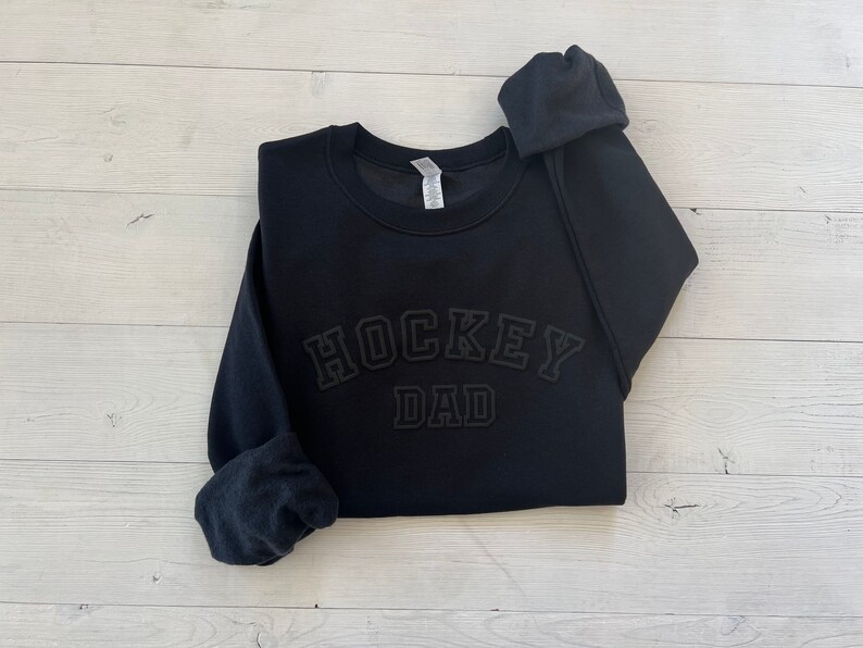 K&ouml;nnte beinhalten: Schwarzer Pullover mit dem Text "HOCKEY DAD" in schwarzer, erhabener Blockschrift.