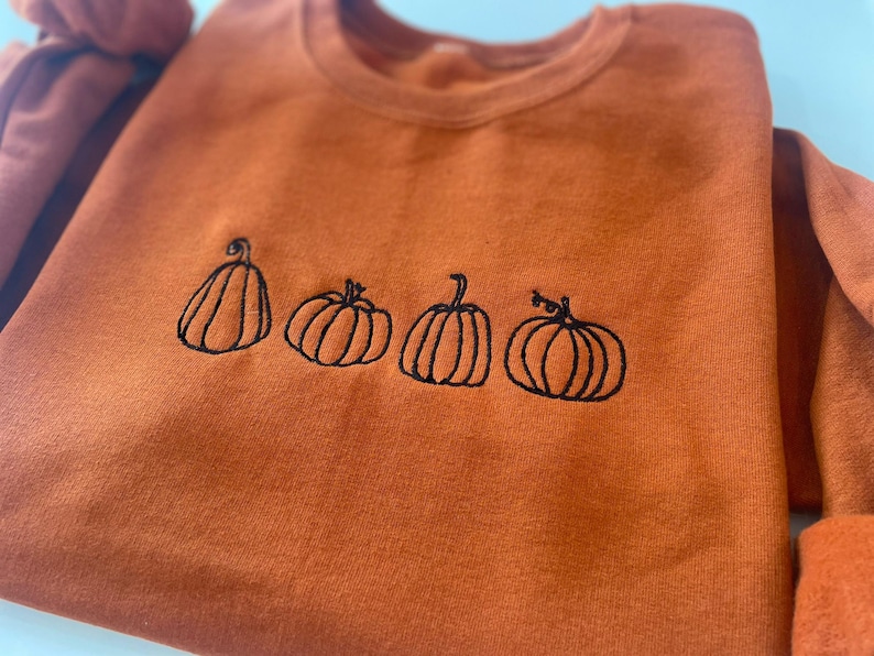 Puede incluir: Una sudadera de color naranja quemado con calabazas bordadas en negro. Las calabazas est&aacute;n en fila y todas son del mismo tama&ntilde;o.