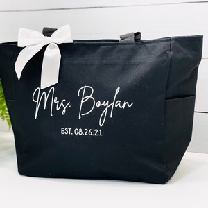 K&ouml;nnte beinhalten: Schwarze Canvas-Tragetasche mit wei&szlig;en Henkeln und einer wei&szlig;en Schleife. Die Tasche hat eine wei&szlig;e Aufschrift mit dem Text "Mrs. Boylan EST. 08.26.21".