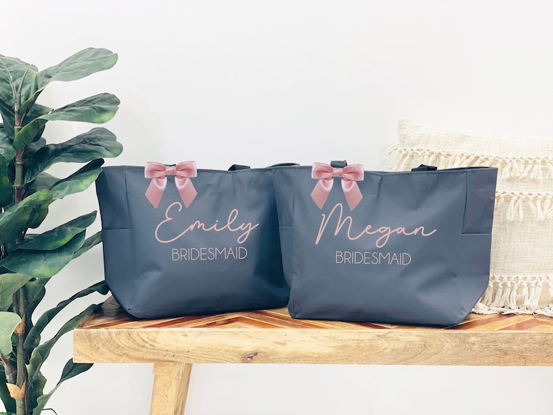 K&ouml;nnte beinhalten: Zwei graue Tragetaschen mit rosa Schleifen und den Namen "Emily Bridesmaid" und "Megan Bridesmaid" darauf gedruckt.