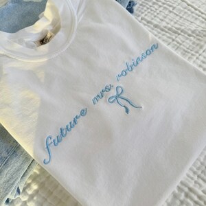 Op de afbeelding: Wit T-shirt met blauwe borduurwerk dat "future mrs. robinson" zegt met een blauwe strik.