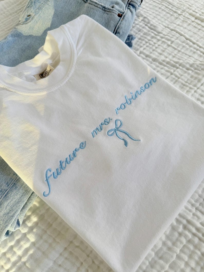 Op de afbeelding: Wit T-shirt met blauwe borduurwerk dat "future mrs. robinson" zegt met een blauwe strik.