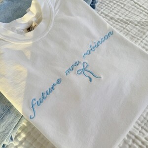 Op de afbeelding: Wit T-shirt met blauwe borduurwerk dat "future mrs. robinson" zegt met een blauwe strik.