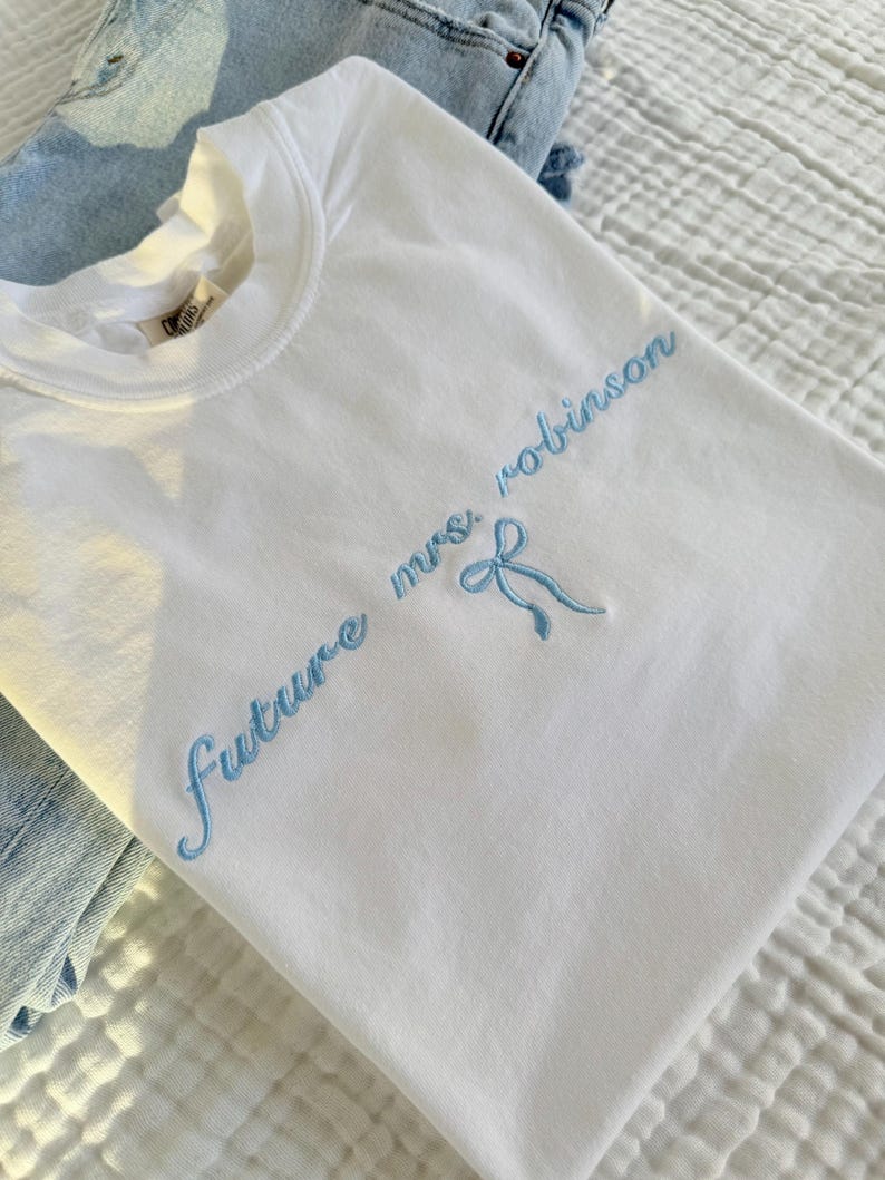 Op de afbeelding: Wit T-shirt met blauwe borduurwerk dat "future mrs. robinson" zegt met een blauwe strik.