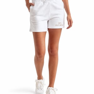 May include: White athletic shorts featuring an elastic waistband, side pockets, and the text EST. 08.17.2026 embroidered in a light tan color on the lower left leg. The shorts are paired with white sneakers that have decorative white embellishments.