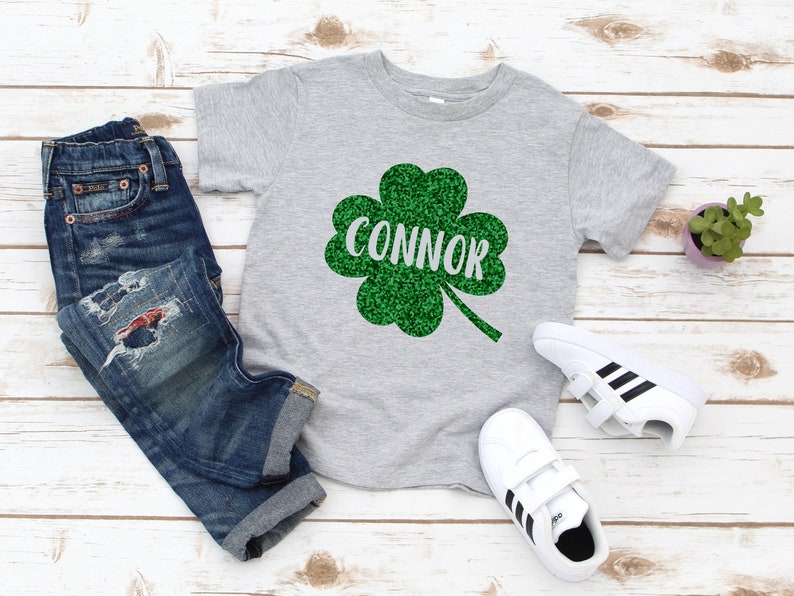May include: A grey t-shirt featuring a green glitter shamrock with the name "CONNOR" in white lettering. The shirt is styled with blue jeans and white trainers with black stripes.