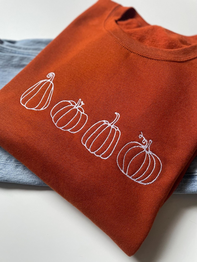 Puede incluir: Una sudadera de color &oacute;xido con un dise&ntilde;o bordado blanco de cuatro calabazas.