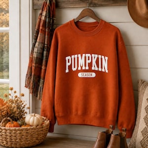Op de afbeelding: Een roestoranje sweater met ronde hals en de witte tekst PUMPKIN SEASON in een collegiaal lettertype op de borst. De trui met lange mouwen heeft een geribbelde halslijn, boorden en zoom voor een klassieke pasvorm.