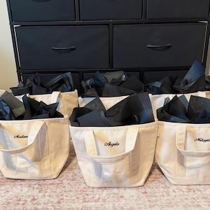 May include: A set of three off-white canvas tote bags, each featuring two handles and black tissue paper tucked inside. The bags are personalized with embroidered names in black script, specifically reading Madison, Angela, and Mikayla.