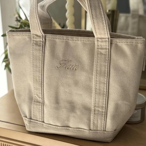 May include: A small, off-white canvas tote bag featuring two sturdy handles and a reinforced base. The front of the bag is embroidered with the name Kate in a cursive script font, centered between the two vertical fabric straps on the exterior.