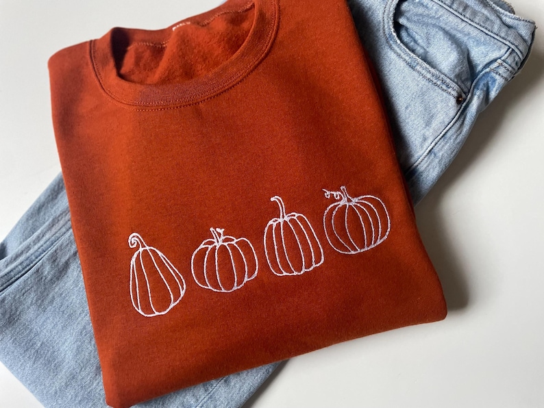 Puede incluir: Una sudadera de color &oacute;xido con un dise&ntilde;o bordado en blanco de cuatro calabazas. Las calabazas est&aacute;n delineadas en blanco y tienen un dise&ntilde;o simple y minimalista.