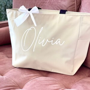 K&ouml;nnte beinhalten: Eine beige Canvas-Tragetasche mit einer wei&szlig;en Schleife und dem Namen "Olivia" in wei&szlig; auf der Vorderseite.