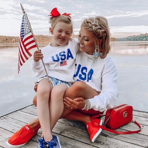 Puede incluir: Una madre y su hija llevan sudaderas blancas con la palabra "USA" impresa en ellas. La hija sostiene una peque&ntilde;a bandera estadounidense. Est&aacute;n sentadas en un muelle de madera junto a un lago. La madre lleva zapatillas rojas y un bolso rojo con una cadena dorada. La hija lleva zapatillas azules.