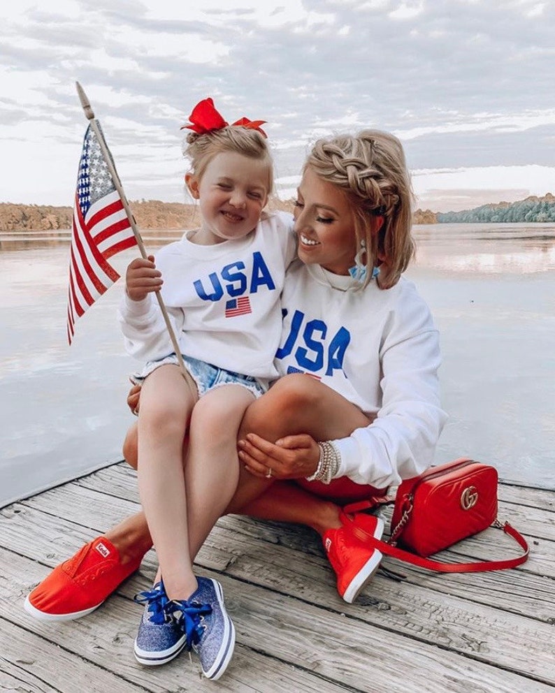 Puede incluir: Una madre y su hija llevan sudaderas blancas con la palabra "USA" impresa en ellas. La hija sostiene una peque&ntilde;a bandera estadounidense. Est&aacute;n sentadas en un muelle de madera junto a un lago. La madre lleva zapatillas rojas y un bolso rojo con una cadena dorada. La hija lleva zapatillas azules.