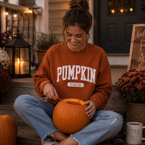 Op de afbeelding: Een roestkleurig sweatshirt met lange mouwen en ronde hals met de witte tekst PUMPKIN SEASON op de borst. Het kledingstuk wordt gecombineerd met een lichtblauwe spijkerbroek met een gerafelde zoom en witte canvas sneakers.
