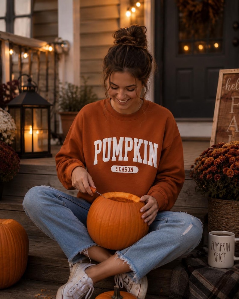 Op de afbeelding: Een roestkleurig sweatshirt met lange mouwen en ronde hals met de witte tekst PUMPKIN SEASON op de borst. Het kledingstuk wordt gecombineerd met een lichtblauwe spijkerbroek met een gerafelde zoom en witte canvas sneakers.