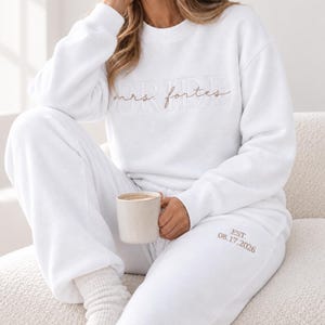 May include: A white crewneck sweatshirt and matching sweatpants set. The sweatshirt features the word BRIDE in white embroidery with the text mrs. fortes in brown script layered over it. The sweatpants feature the text EST. 08.17.2026 in brown.