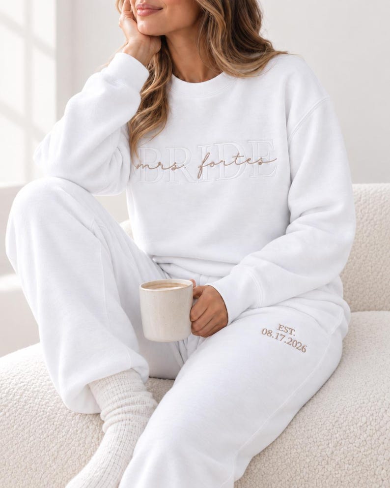 May include: A white crewneck sweatshirt and matching sweatpants set. The sweatshirt features the word BRIDE in white embroidery with the text mrs. fortes in brown script layered over it. The sweatpants feature the text EST. 08.17.2026 in brown.