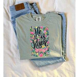 May include: A light green t-shirt with a floral design and the text "He is Risen" with the scripture reference "Luke 24:6-7".