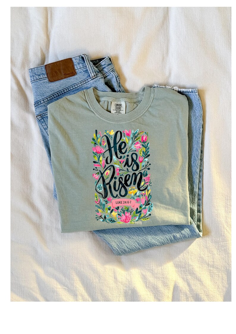 May include: A light green t-shirt with a floral design and the text "He is Risen" with the scripture reference "Luke 24:6-7".