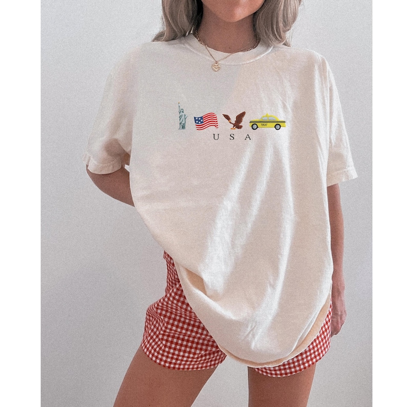 Puede incluir: Camiseta color crema con un gr&aacute;fico de la Estatua de la Libertad, una bandera estadounidense ondeando, un &aacute;guila y un taxi amarillo. La palabra "USA" est&aacute; impresa debajo. La persona lleva unos shorts a cuadros rojos y blancos.