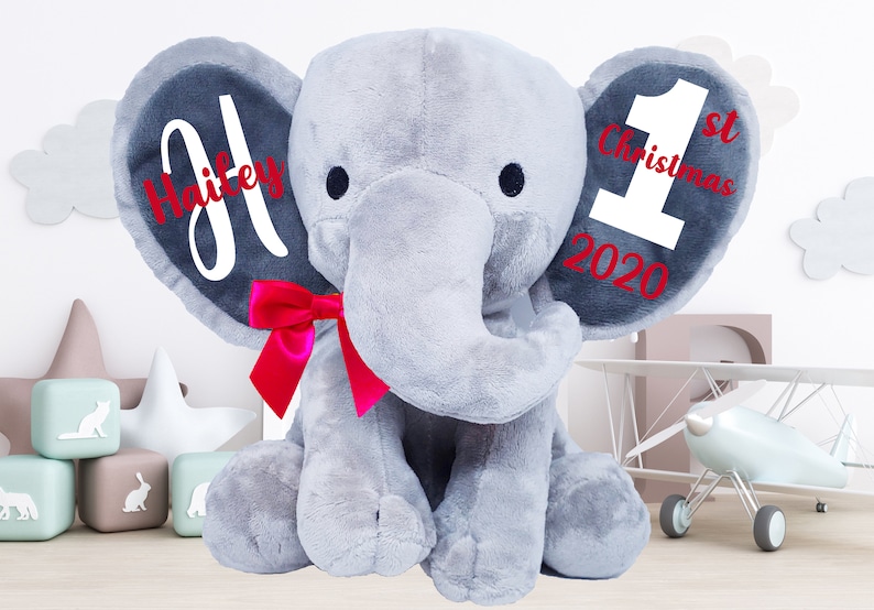 May include: A gray plush elephant with a red bow. The elephant's ears are personalized with the name "Hailey" and the text "1st Christmas 2020".