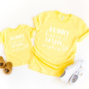 May include: Two yellow t-shirts with the text "Bunny kisses & Easter wishes" in white. The larger shirt has a larger text design. The smaller shirt has a smaller text design.