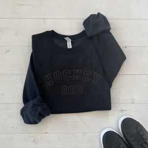 K&ouml;nnte beinhalten: Schwarzer Pullover mit dem Text "HOCKEY DAD" in schwarzer, erhabener, umrahmter Schrift.