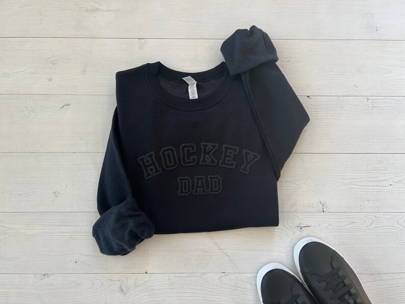 K&ouml;nnte beinhalten: Schwarzer Pullover mit dem Text "HOCKEY DAD" in schwarzer, erhabener, umrahmter Schrift.