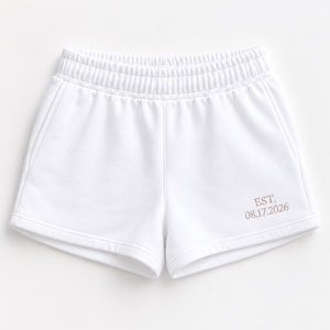 May include: White athletic shorts featuring an elastic waistband and two side pockets. The lower right leg displays embroidered text in a light tan color that reads EST. 08.17.2026. The shorts are constructed from a smooth, solid white fabric.