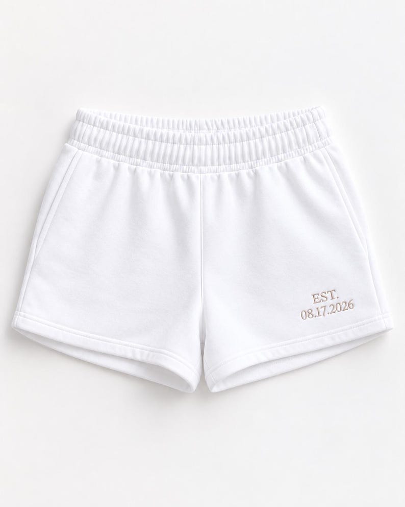 May include: White athletic shorts featuring an elastic waistband and two side pockets. The lower right leg displays embroidered text in a light tan color that reads EST. 08.17.2026. The shorts are constructed from a smooth, solid white fabric.