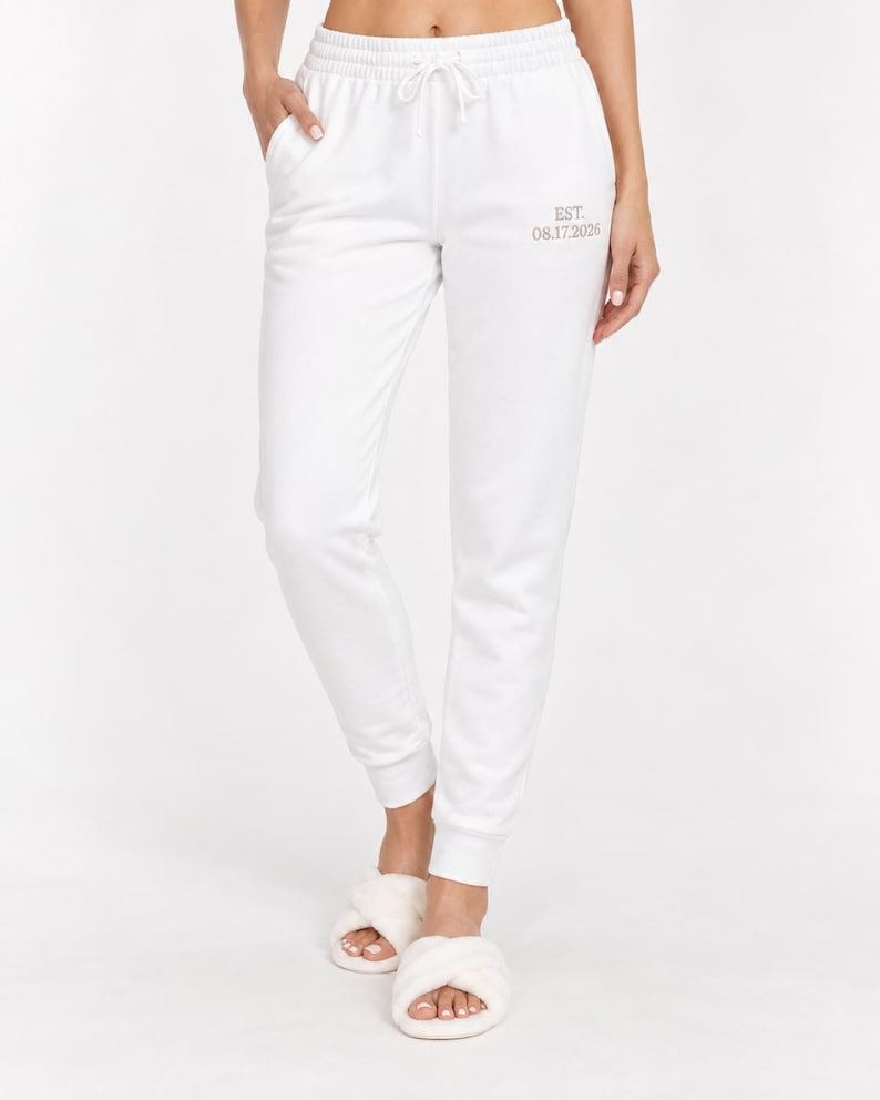 May include: White sweatpants featuring an elastic waistband with a drawstring and side pockets. The left thigh area displays the text EST. 08.17.2026 in a small, light-colored font. The pants have a tapered leg design with ribbed cuffs at the ankles.
