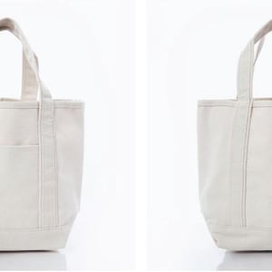 May include: A cream-colored canvas tote bag featuring two sturdy top handles, a flat base, and a single exterior slip pocket on the front. The bag is constructed from a durable woven fabric with reinforced stitching along the handles and seams.