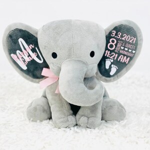 May include: A gray plush elephant with a pink bow and black eyes. The elephant's ears have custom text and baby details: date, time, weight, and footprints. The text on the left ear says "Makenna Grace".