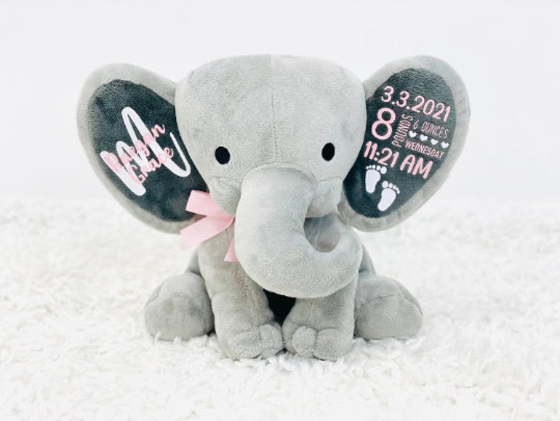 May include: A gray plush elephant with a pink bow and black eyes. The elephant's ears have custom text and baby details: date, time, weight, and footprints. The text on the left ear says "Makenna Grace".
