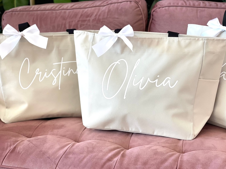 K&ouml;nnte beinhalten: Drei beigefarbene Canvas-Tragetaschen mit wei&szlig;en Schleifen und schwarzen Griffen. Die Taschen sind mit den Namen "Cristina", "Olivia" und "A" in wei&szlig;er Schrift personalisiert.