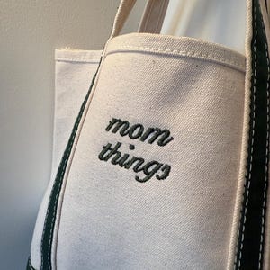 May include: Off-white canvas tote bag with the words "mom things" embroidered in black. The bag has a rectangular shape with a reinforced green base and green trim along the edges. The handles are a natural beige color.
