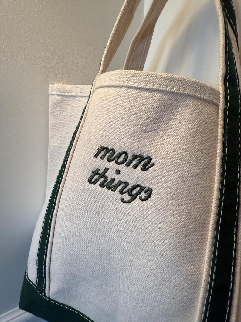 May include: Off-white canvas tote bag with the words "mom things" embroidered in black. The bag has a rectangular shape with a reinforced green base and green trim along the edges. The handles are a natural beige color.