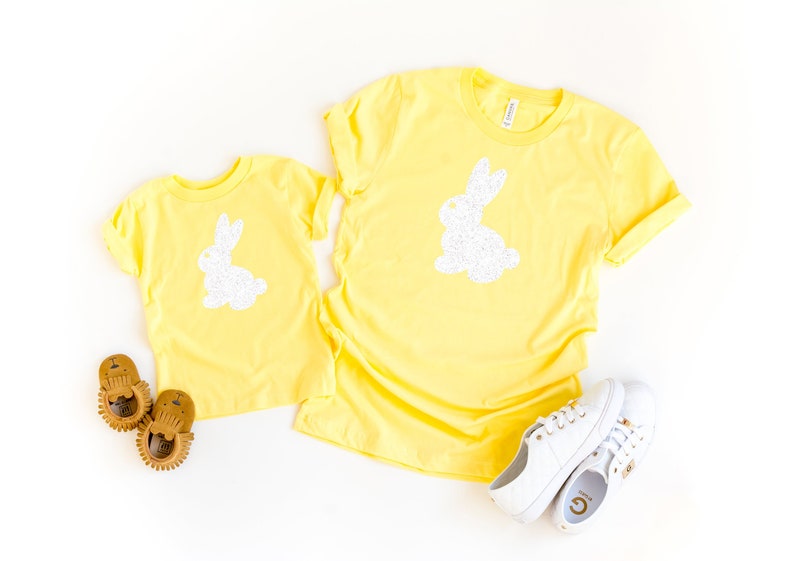 May include: Two yellow t-shirts, one adult and one child size, each featuring a white glitter bunny design. White sneakers and brown baby shoes are also in the image. The shirts are likely for Easter or spring.