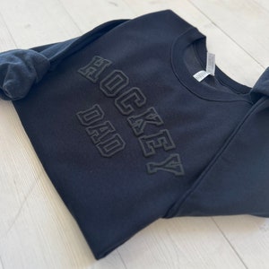 K&ouml;nnte beinhalten: Schwarzer Pullover mit dem Schriftzug "Hockey Dad" in erhabener Schrift.