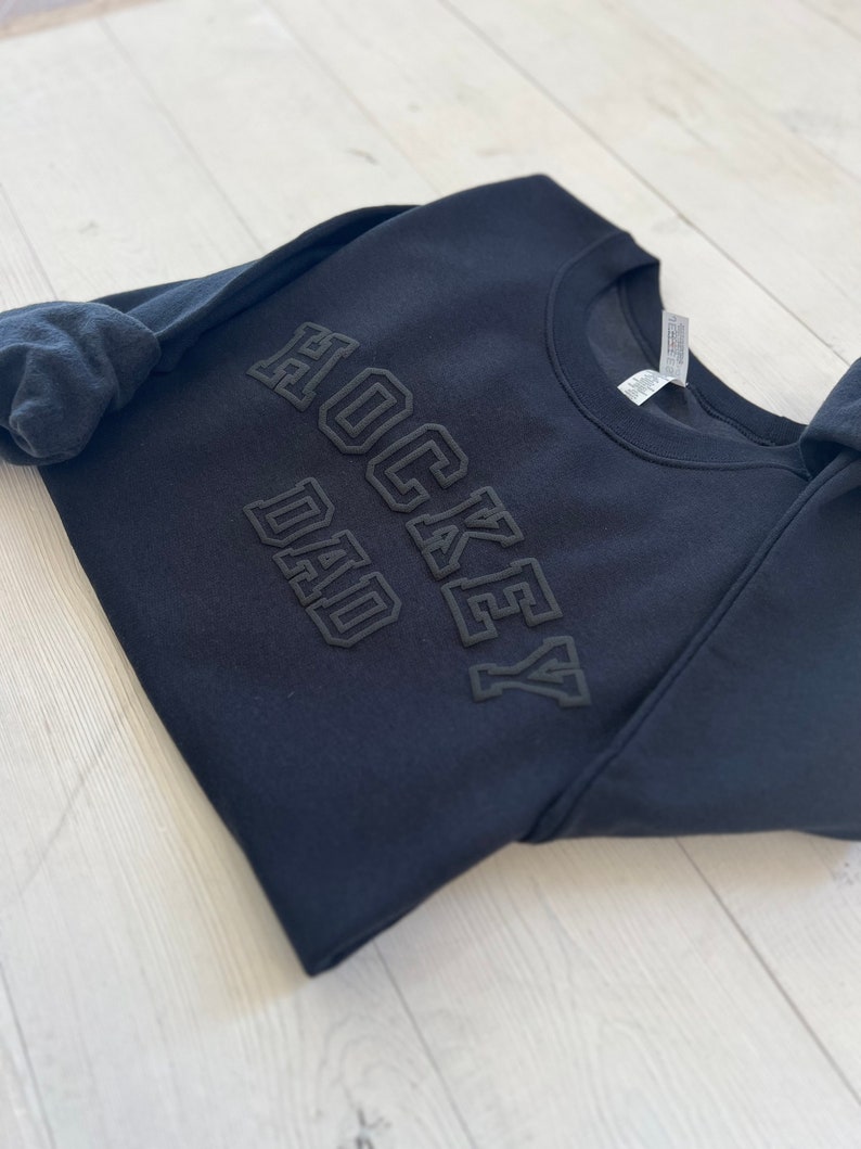 K&ouml;nnte beinhalten: Schwarzer Pullover mit dem Schriftzug "Hockey Dad" in erhabener Schrift.